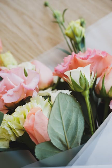 A close-up of an elegant bouquet featuring pale pink and cream lisianthus flowers with ruffled petals, complemented by fresh green leaves and stems. The bouquet is partially wrapped in translucent whi