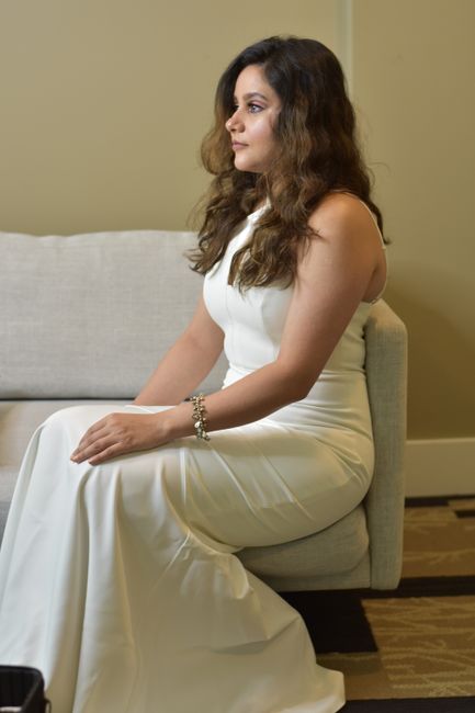 A woman with long, wavy brown hair wearing a white sleeveless dress is seated on a light grey upholstered sofa in an indoor setting with soft, neutral-colored walls. Her hands rest on her lap, and she