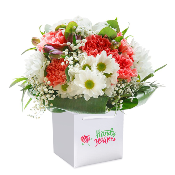 Presented in a pristine, white square gift bag, this exuberant floral arrangement radiates freshness and affection, perfectly embodying the uplifting character of Surrey Quays. An artful mix of ruffled coral and peach carnations fills the bouquet with lively colour and sumptuous texture, each blossom brimming with subtle fragrance. White daisies, their petals crisp and fine, are distinguished by bright lime-green centers that shimmer beneath the soft indoor lighting, reminiscent of riverside meadows not far from Canada Water. Dainty clusters of baby's breath weave through the composition, imparting an ethereal lightness and accentuating the bouquet's round, harmonious silhouette. On the left edge, unopened buds tinged with purple and pink offer quiet contrast and a sense of unfolding beauty. The ensemble is embraced by a generous layer of lustrous, dark green foliage-broad, heart-shaped leaves providing a natural, elegant frame for the display. The finely finished white gift bag, with sturdy ribbon handles at the top, promises easy delivery to addresses from Lower Road to the wharf side apartments. This evocative presentation blends craftsmanship, elegance, and a touch of local flair, making it ideal for gifting on birthdays, anniversaries, or gestures of gratitude within Surrey Quays' thriving waterside community.