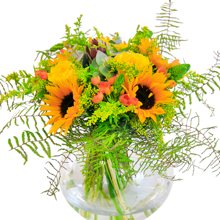 The first impression this Surrey Quays bouquet delivers is one of golden exuberance and joyful artistry, echoing the area's vibrant local spirit. Sunflowers in full flourish dominate the composition, their radiant yellow petals and textured, chocolate-brown centers reminiscent of late summer days in King Stairs Gardens or along the canals. Supporting their brightness are plump yellow garden roses, each bloom a spiral of dense petals unfurling into luminous rosettes. Scattered amongst these are clusters of glossy red Hypericum berries, their ruby hue gleaming like hidden gemstones and providing a visual spark that sets off the yellows and greens. Cascading ribbons of feathery green fern layers add movement, their softness balancing the strong form of the sunflowers, while light green sprigs of goldenrod with miniature yellow florets brighten the gaps with a sense of wild meadow charm. The inclusion of slender green spikes and subtle blue-green eucalyptus foliage heightens the bouquet's texture and depth, infusing the fragrant arrangements seen across Surrey Quays' local homes and markets. All these blooms are anchored in a spherical, transparent glass vase-clean lines cradling crisscrossed stems immersed in water, lending freshness and clarity that enhances the overall display. This arrangement is not just a feast for the eyes and senses; it's a thoughtful choice for milestones or spontaneous celebrations, suffusing living rooms and gathering spaces in Surrey Quays with happiness and the promise of new memories.